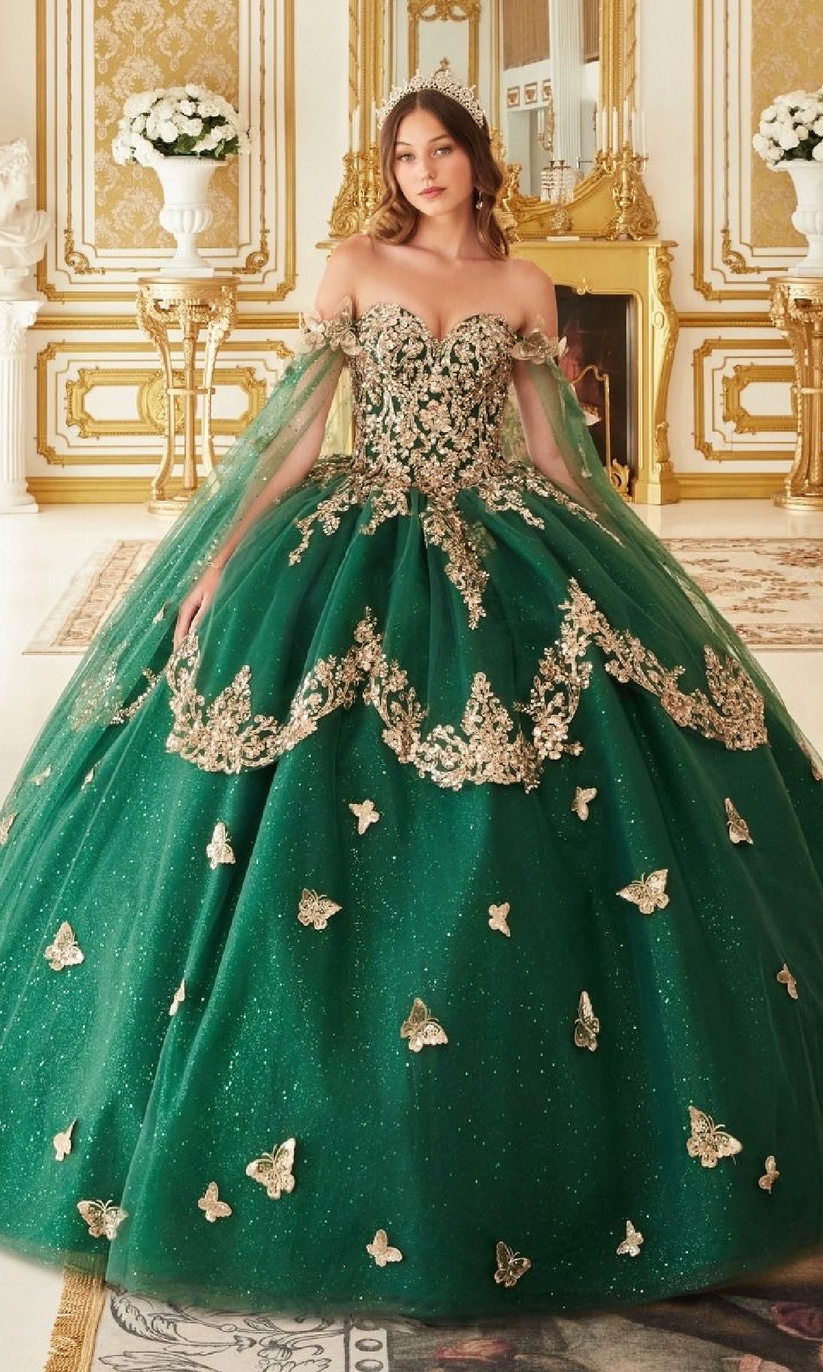 A woman wearing the Quinceanera Dress 15721 by Ladivine—emerald green with gold embroidery and butterfly accents—stands in a lavish room decorated with gold details and floral arrangements.